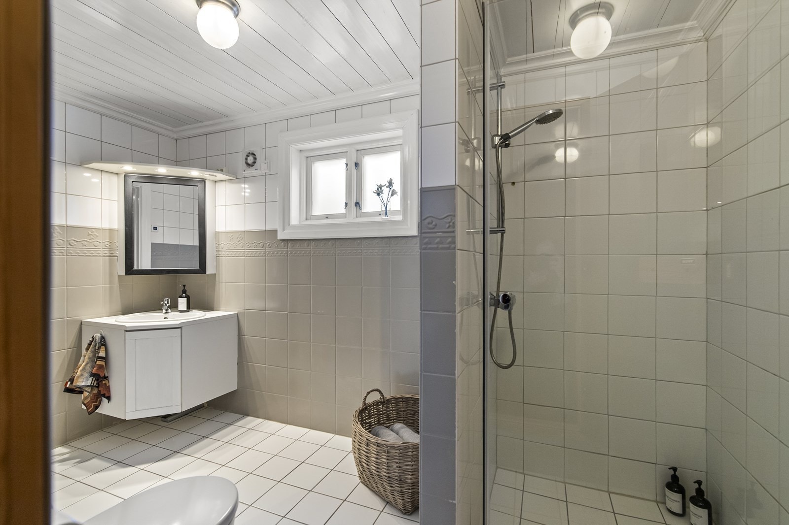 Badet i kjelleretasjen er innredet med grå og hvite fliser, og har toalett, gulvvarme og åpen dusjnisje. Et hvitt servantskap har tilhørende speilskap med belysning, og tilliggende badet finner man en badstue med plassbygde trebenker. Galleribilde