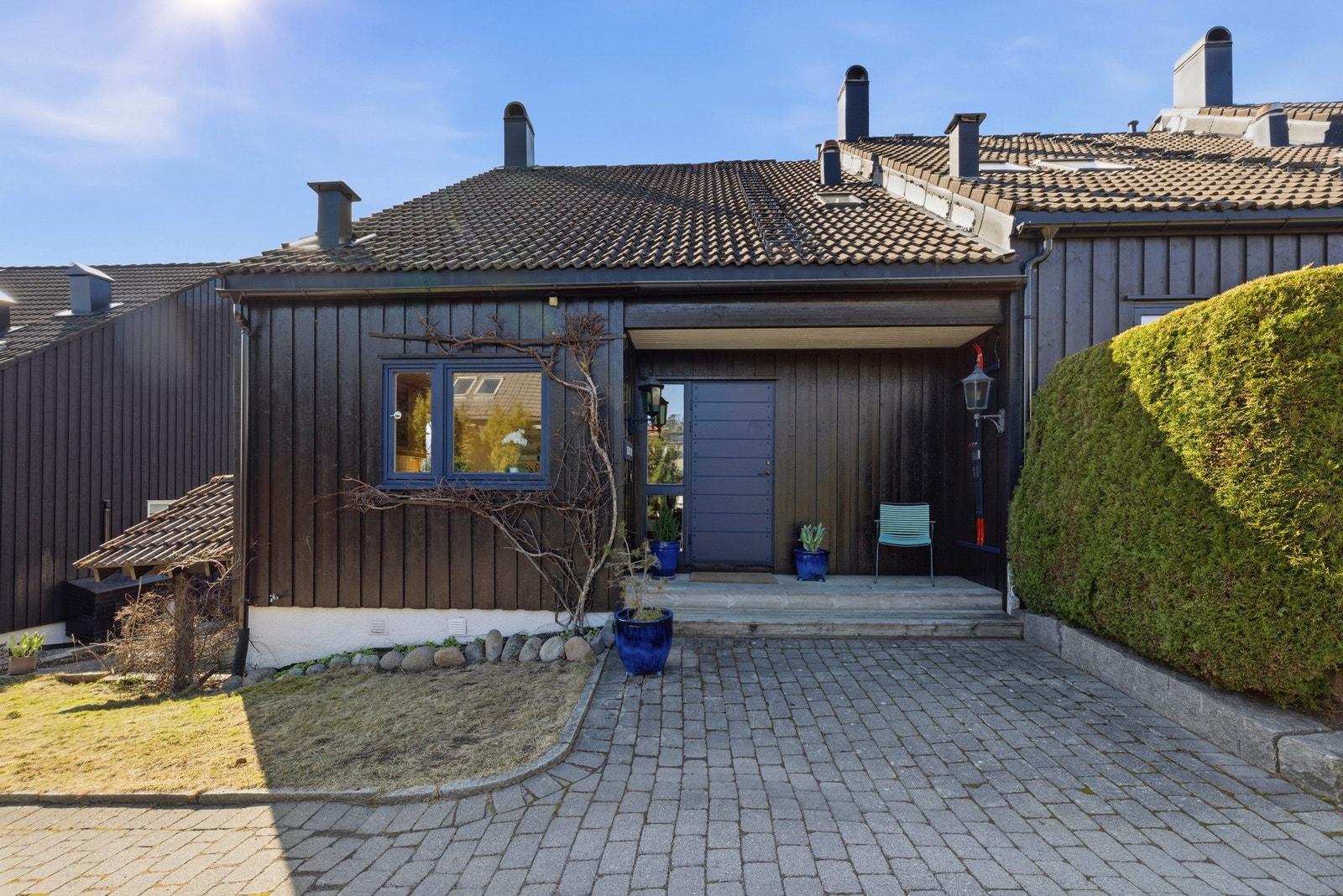 Representativt inngangsparti med brostein og gressplen og skistativ på veggen utenfor døren. Det er også inngang til vaskerom i kjelleren, på sydsiden av huset. Galleribilde