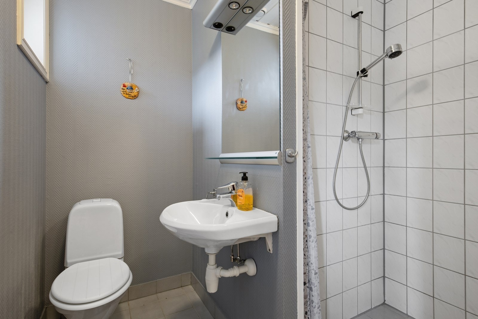Badet i første etasje har flislagt gulv med gulvvarme og er innredet med vegghengt servant, gulvstående toalett og dusjnisje med forheng. Badet har et visst oppgraderingsbehov, noe som gir ny eier mulighet til å sette sitt eget preg. Galleribilde