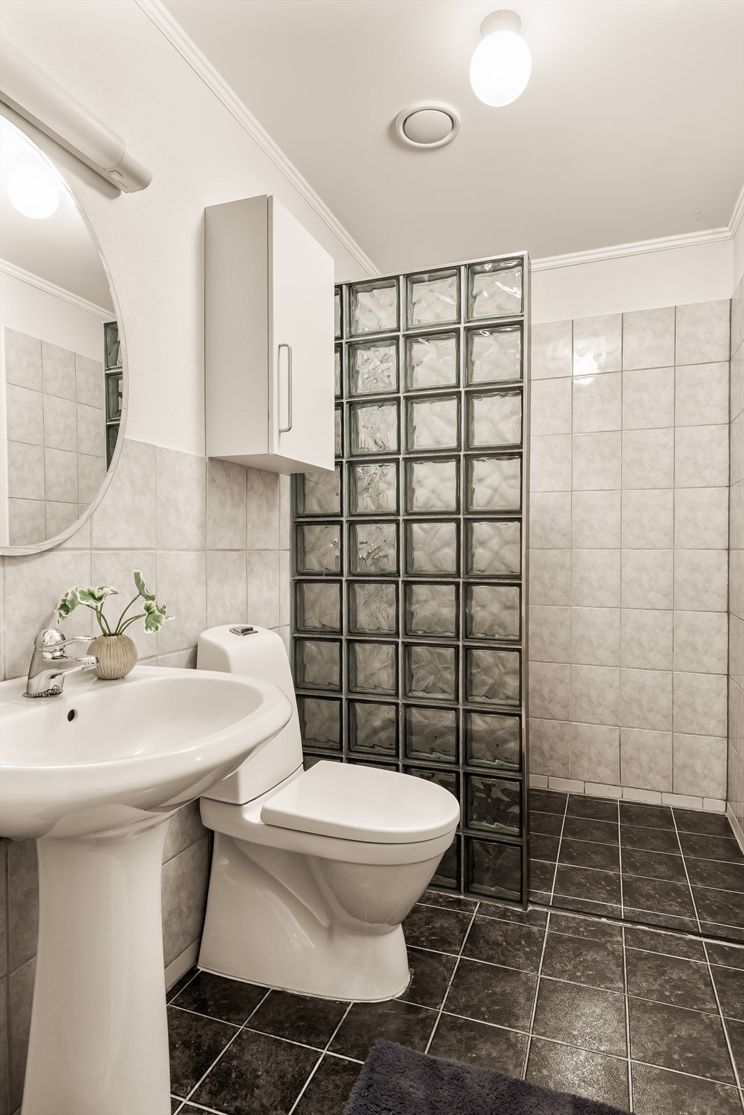 Gulvet er belagt med fliser og har varmekabler. Badet er innredet med toalett, søyleservant, speil og veggskap som gir gode oppbevaringsmuligheter.
Dusjsonen er utformet som en egen nisje med vegg i glassbyggerstein. Galleribilde