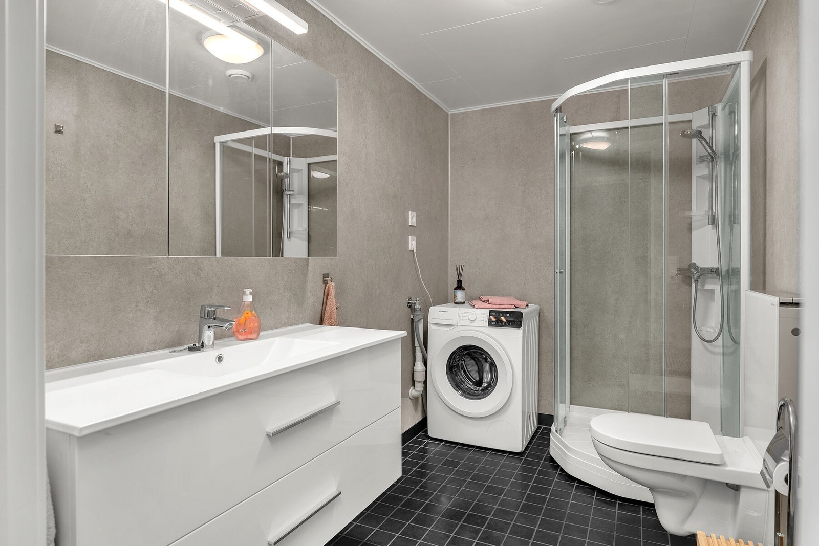 Badet har varmekabler i gulv, opplegg for vaskemaskin/tørketrommel, servant på skuffeseksjon, speilskap, veggmontert toalett og dusjkabinett. Galleribilde