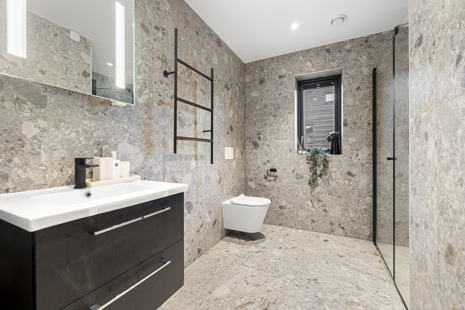 Baderom i moderne utførelse og med lekre fliser. Badet innehar vegghengt toalett, servant med skuffer og dusj innfelt i nisje i vegg. Galleribilde
