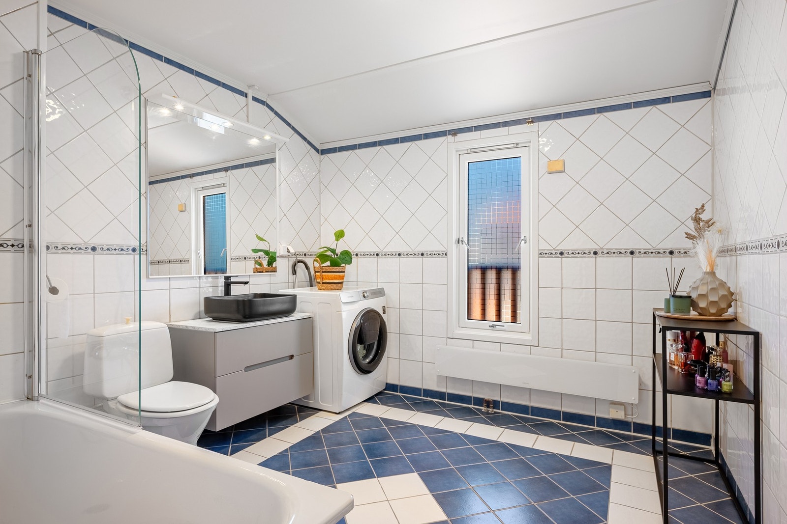 Badet fremstår med flislagte gulv, flislagte vegger og malt tak, og er innredet med servant i møbel, toalett, badekar samt opplegg for vaskemaskin. Galleribilde