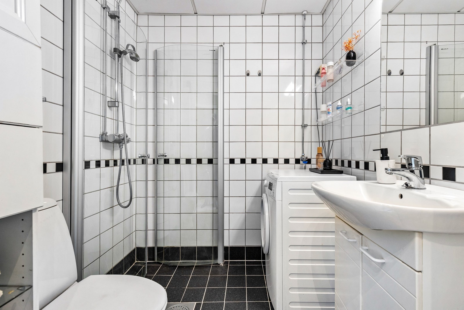 Badet er flislagt på gulv og vegger med elektrisk gulvvarme. Badet er innredet med dusjhjørne med svingbare glassdører, toalett og servant med innredning. Galleribilde