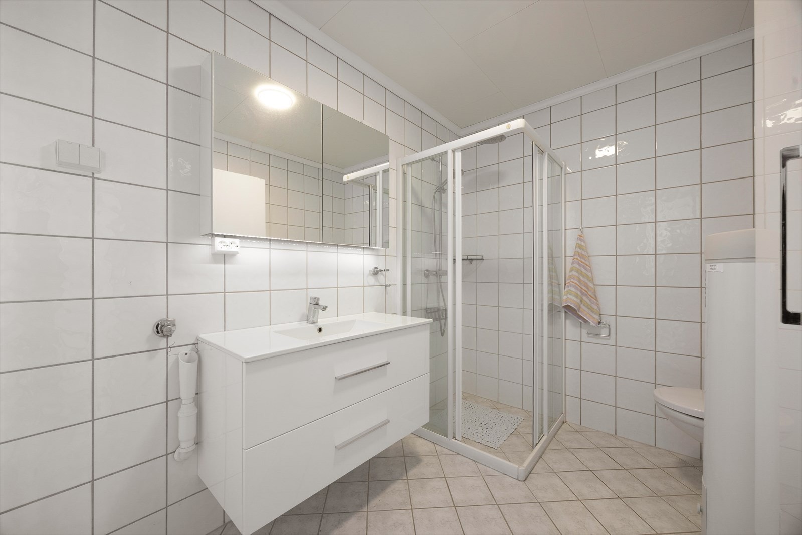 Pent flislagt bad med varmekabler i gulv. Badet inneholder veggmontert toalett, glassdører mot dusjnisje samt dusjgarnityr. Baderoms innredning (2022) med skuffer og heldekkende servant samt
vegghengt skap med speil. Det er også 1.stk vegghengt høyskap. Galleribilde
