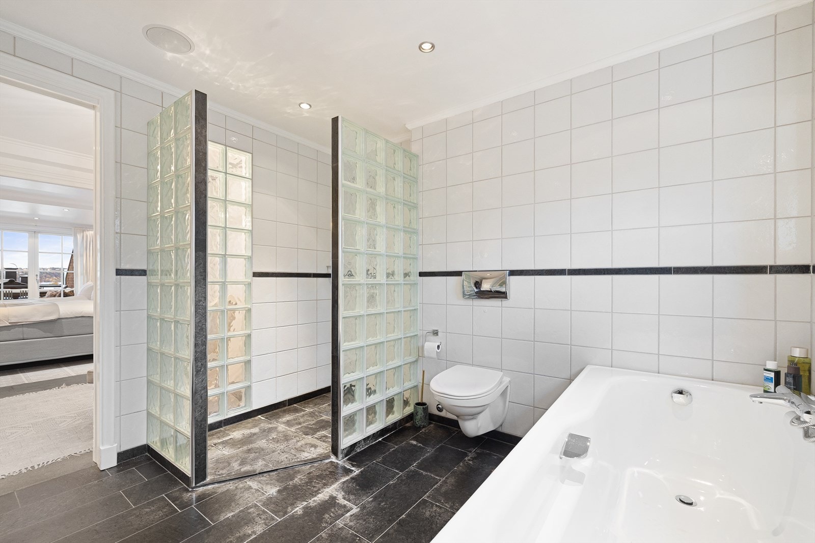 Badet har elektrisk håndkletørker, innfliset badekar, vegghengt toalett og dusjhjørne med glassbyggestein på begge sider. Galleribilde