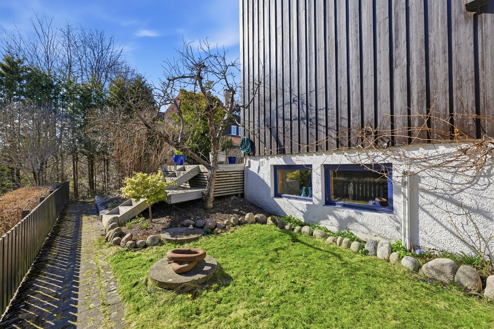Det er enkel adkomst til hagen fra sydenden av huset. Opp langs veggen vokser en drueranke som gir mange og svært gode druer. Legg merke til de store vinduene til kjellerstuen. Galleribilde