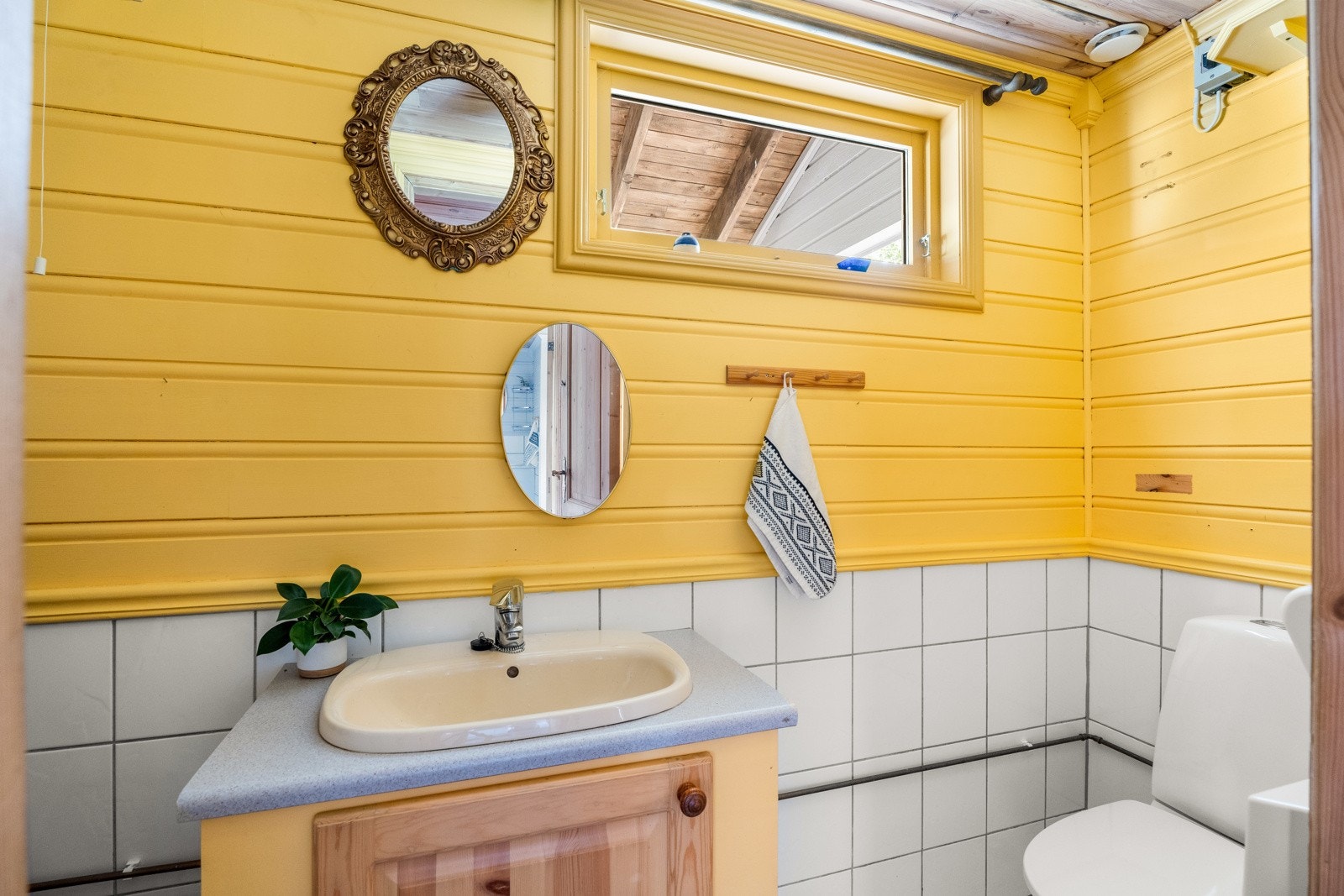 Toalettrommet har fliser på gulv, hvite veggfliser til brystningshøyde, gulmalte panelvegger øverst og et smalt vindu. Galleribilde