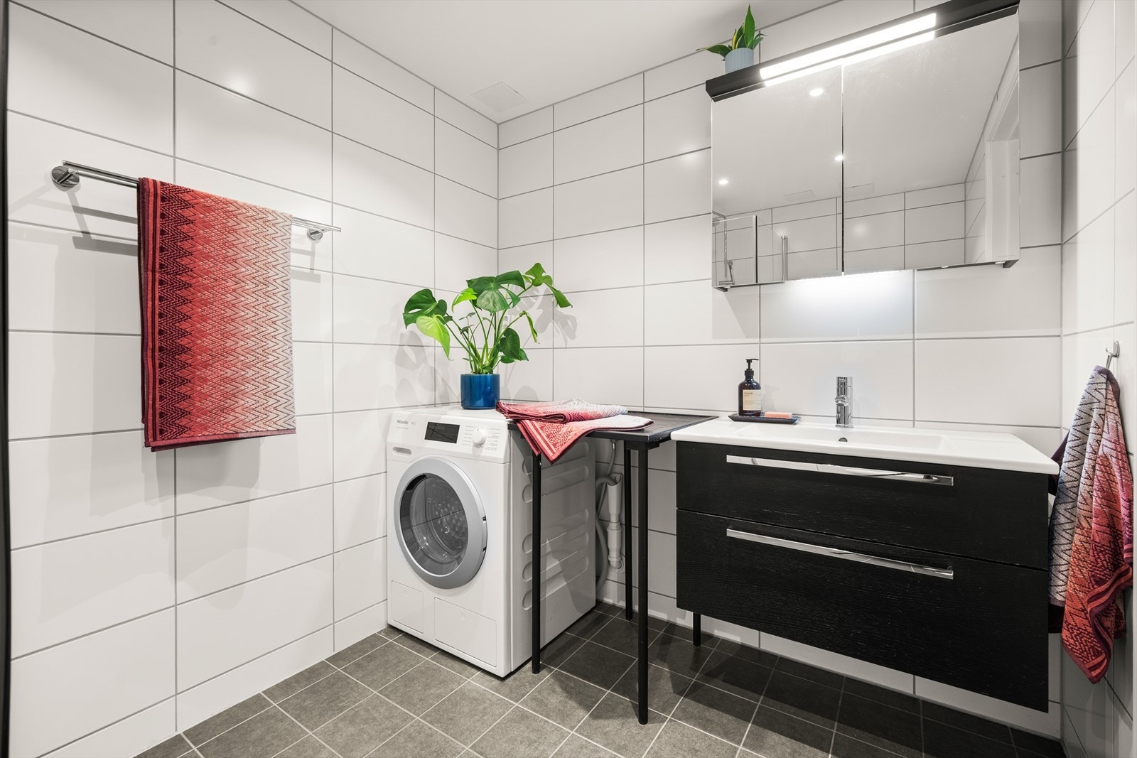 Det er et 80cm bredt servantskap med mørklaminert skuffefronter i spon med aluminiumshåndtak og hvit keramisk enkel servant med ett-greps blandebatterifra Tap well. Speillskap over servanten med lysarmatur over og under skapet. Galleribilde