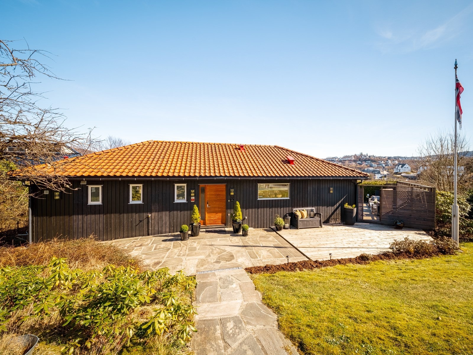Idyllisk opparbeidet tomt med skiferheller ned mot terrasse og inngangsparti, hage med flaggstang og variert beplantning. Det er varmekabler i bakken på vei ned til inngangsdøren fra hovedvei Galleribilde