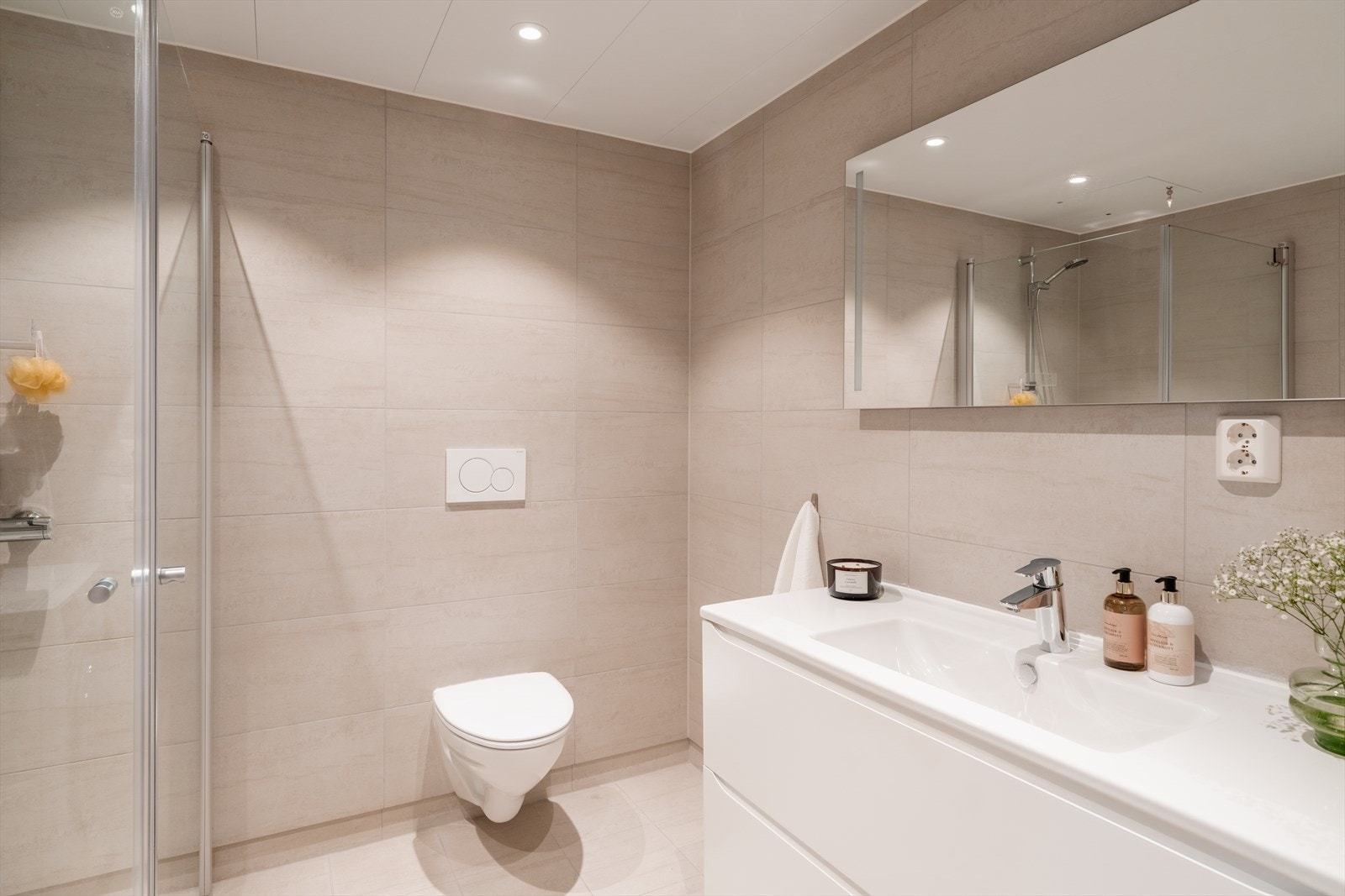 Baderomsinnredningen har hvite fronter, og består av en heldekkende servant på en skuffeseksjon og et stort speil med integrert belysning. Videre har badet et vegghengt toalett, opplegg for vaskemaskin og et dusjhjørne med innfellbare glassdører. Galleribilde