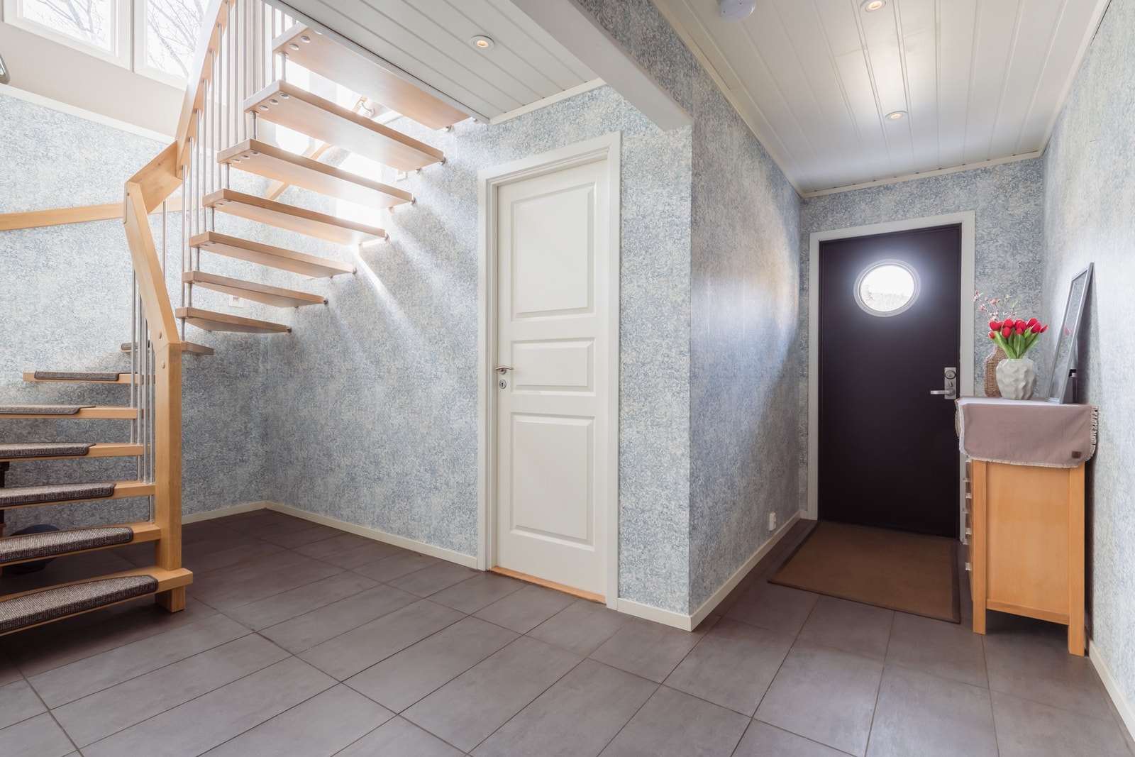 Vel inne er entreen innredet med downlights, grå gulvfliser og mønstret tapet på veggene. Varmekabler i gulv. Galleribilde