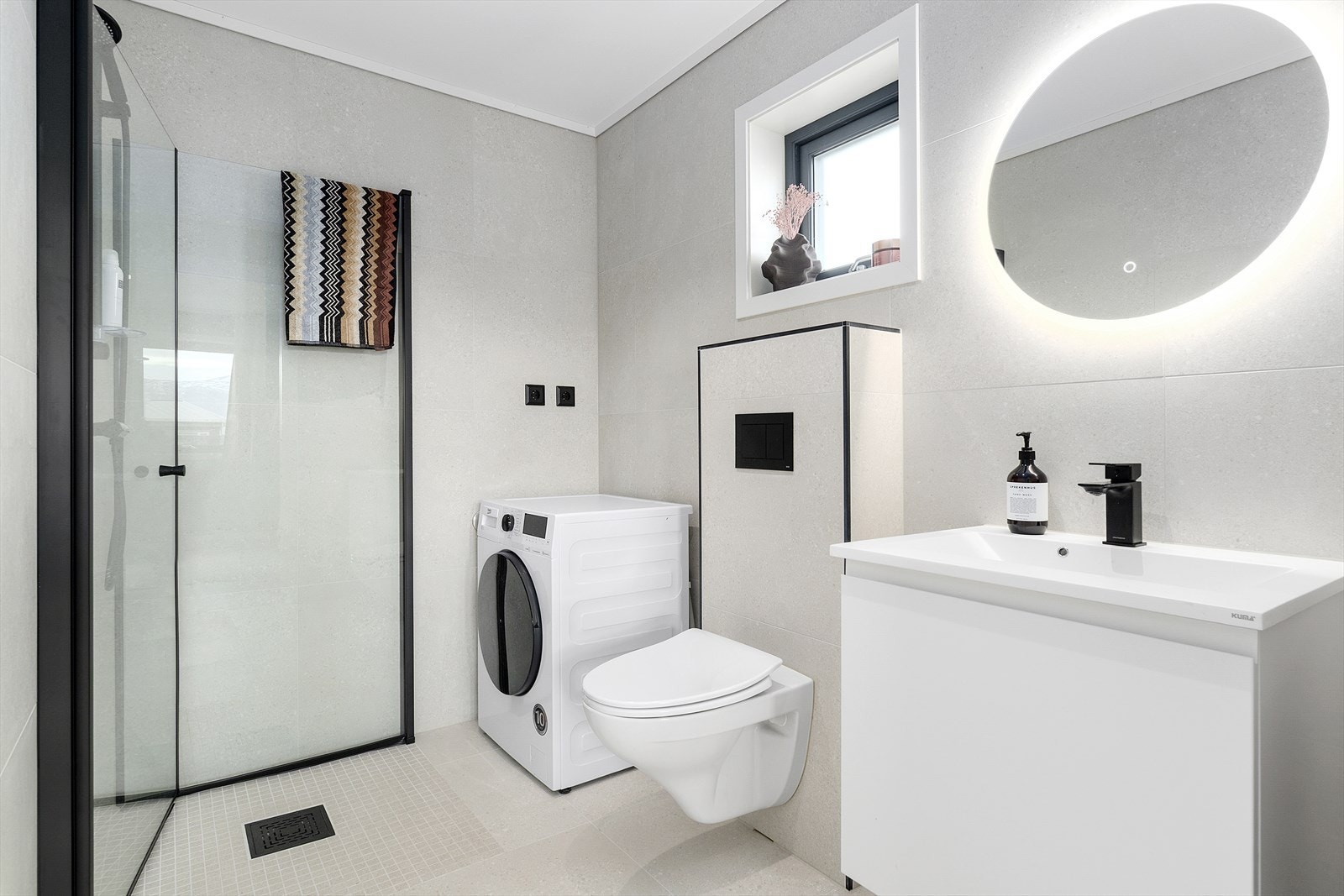 Badet i førsteetasjen har dusjhjørne med innfellbare dører, plass til vaskemaskin og tørketrommel, vegghent wc, servant med oppbevaring og speil med lys. Galleribilde