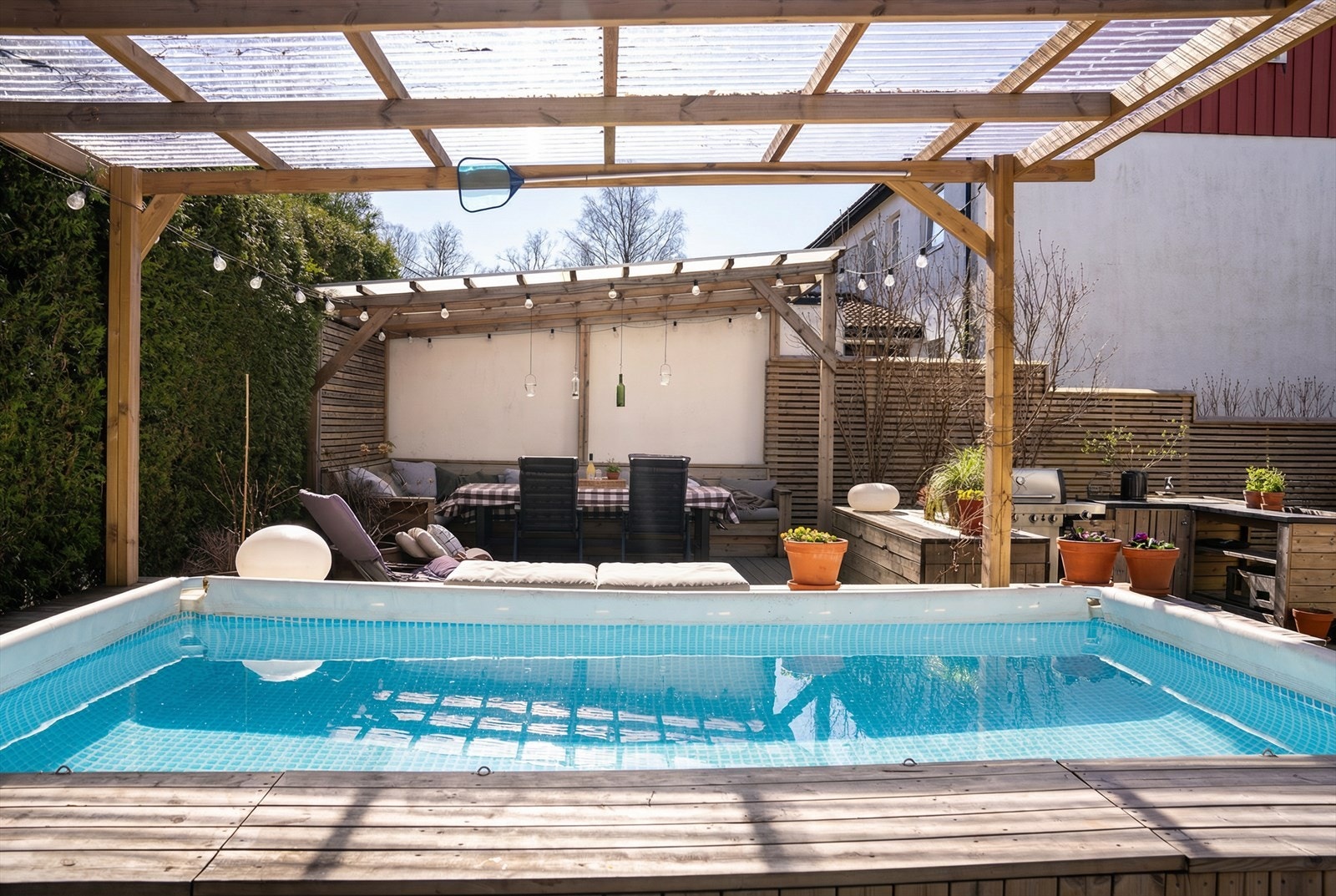 Uteområdet er tilrettelagt med flere sosiale soner, inkludert en pergola med sittegruppe, et utekjøkken og et nedsenket basseng med tredekke rundt. Bildet er AI generert. Galleribilde