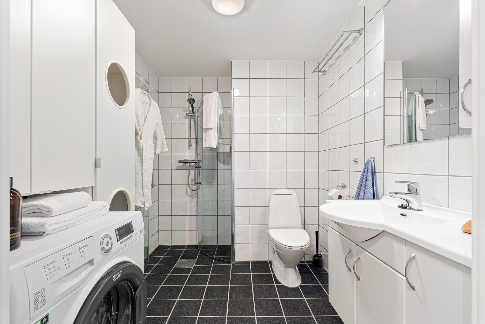 Lyst baderom med dusjkabinett, gulvmontert WC, servant, underskap, speil og veggskap. Galleribilde