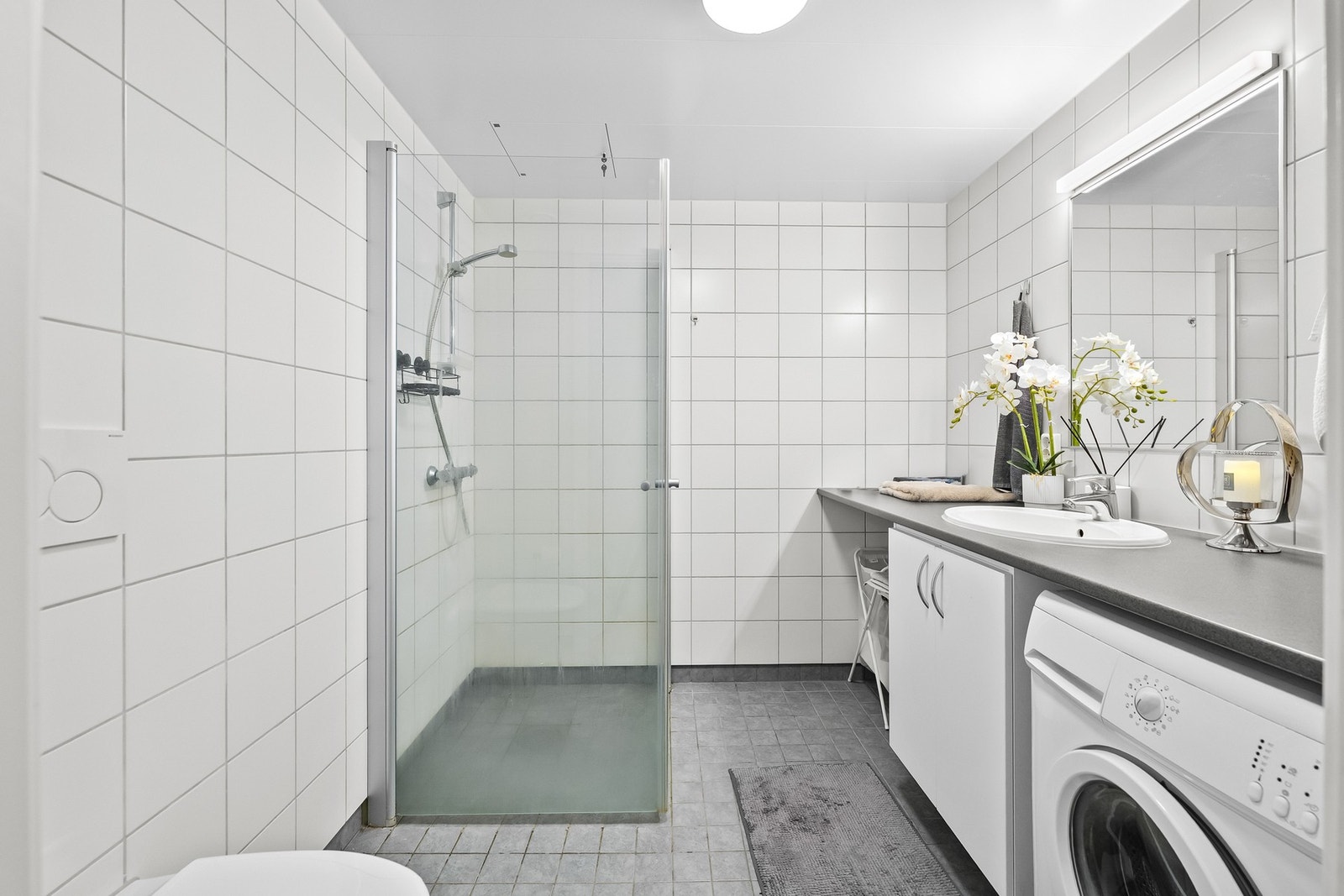 Stilrent bad med flislagte overflater og et moderne uttrykk. Badet er utstyrt med dusjsone, servant med oppbevaring og opplegg for vaskemaskin. Galleribilde