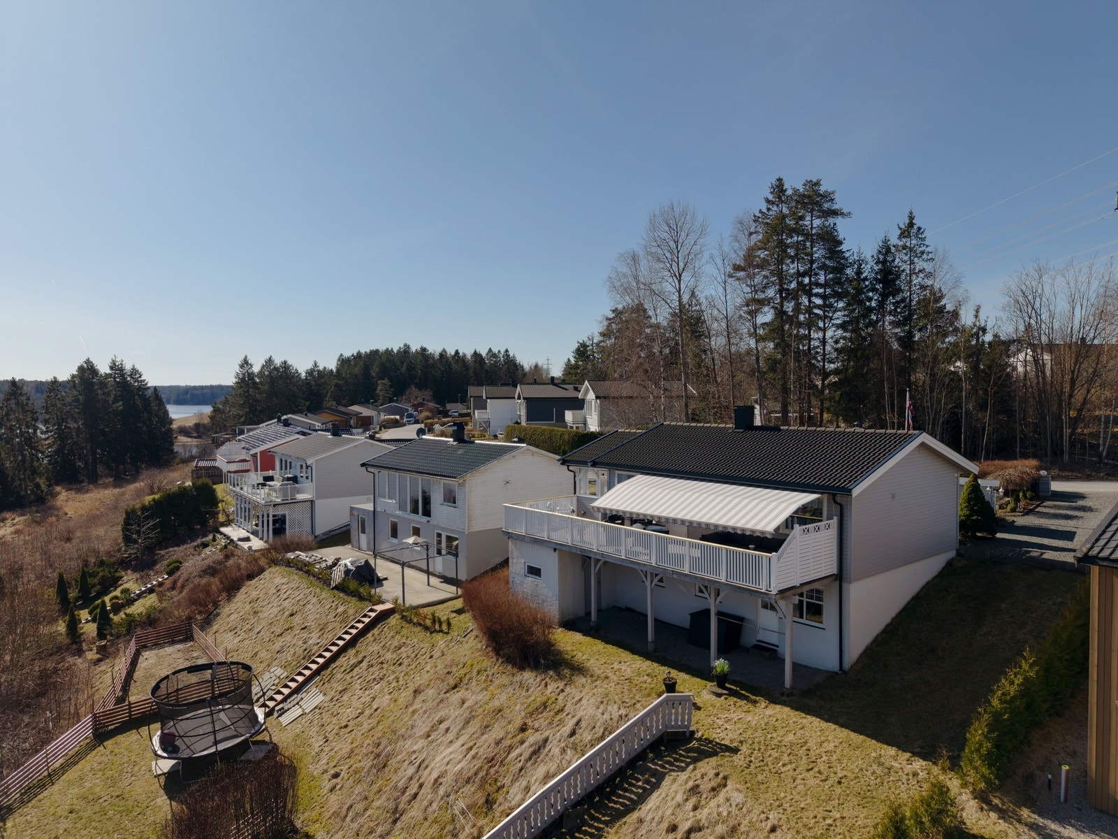 Bilde av fasade og tomt fra øst. Sokkeletasje med utgang til uteplass og bod nede. Hagen skråner noe her og hekk er satt opp i nedre del for å vise tomteskille Galleribilde