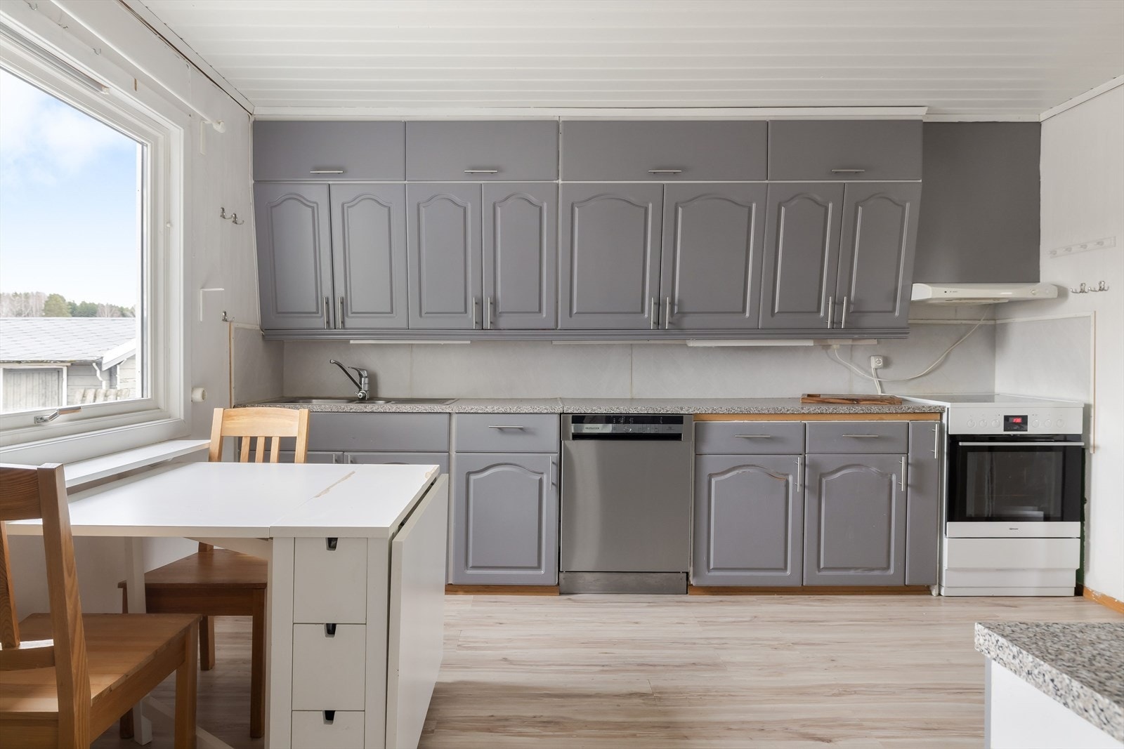 Kjøkkeninnredning med profilerte fronter og benkeplate av laminat. Det er en laminert plate mellom kjøkkenbenk og overskap. Kjøkkenet er utstyrt med ventilator i overskap. Det er frittstående komfyr, oppvaskmaskin og kjøleskap. Galleribilde