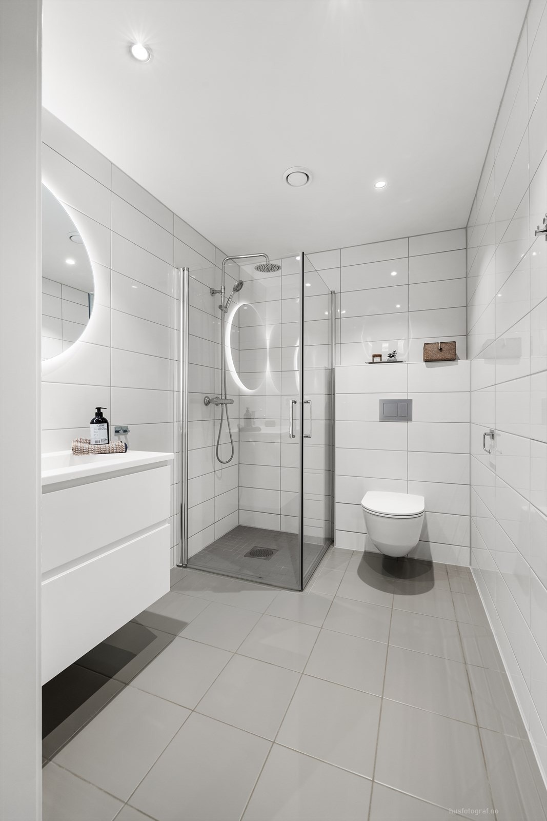 Badet har vegghengt toalett samt dusjhjørne med innfellbare dusjvegger. Det er fliser på gulv og vegger, spotter i himling. Galleribilde