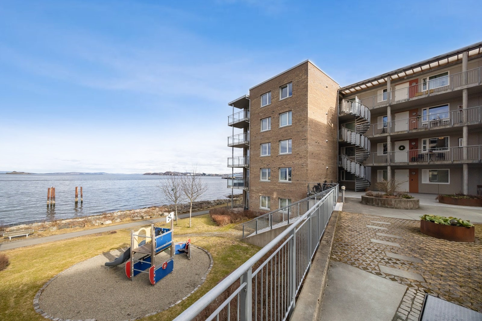 Ettermiddags- og tidlig kveldssol ved inngangspartiet som vender mot en åpen og rolig plass med benker og beplanting. Galleribilde