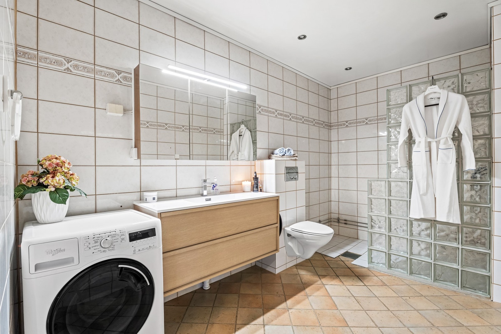 Velholdt og stilrent bad med fliser på veggene og fliser på gulv med elektrisk gulvvarme. Badet er utstyrt med et vegghengt toalett og et dusjhjørne adskilt med vegg av glassbyggerstein. Galleribilde