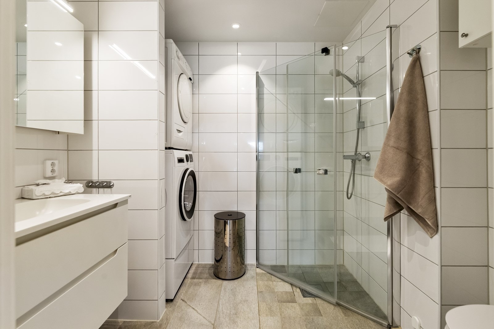 Badet inneholder toalett, dusjhjørne med glassdører, servant i baderomsinnredning med underskuffer og speilskap, samt tilkobling for vaskemaskin og tørketrommel. Galleribilde