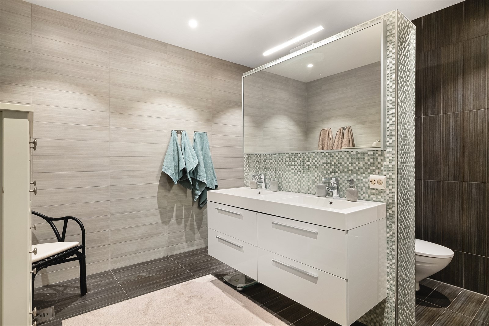 Badet er helfliset og har en moderne standard med delikate fargevalg og praktiske løsninger. En halvvegg med mosaikkfliser deler rommet mellom toalett og dusjnisje, og badet er utstyrt med vegghengt toalett, innredning med nedfelt servant og stort speil. Galleribilde