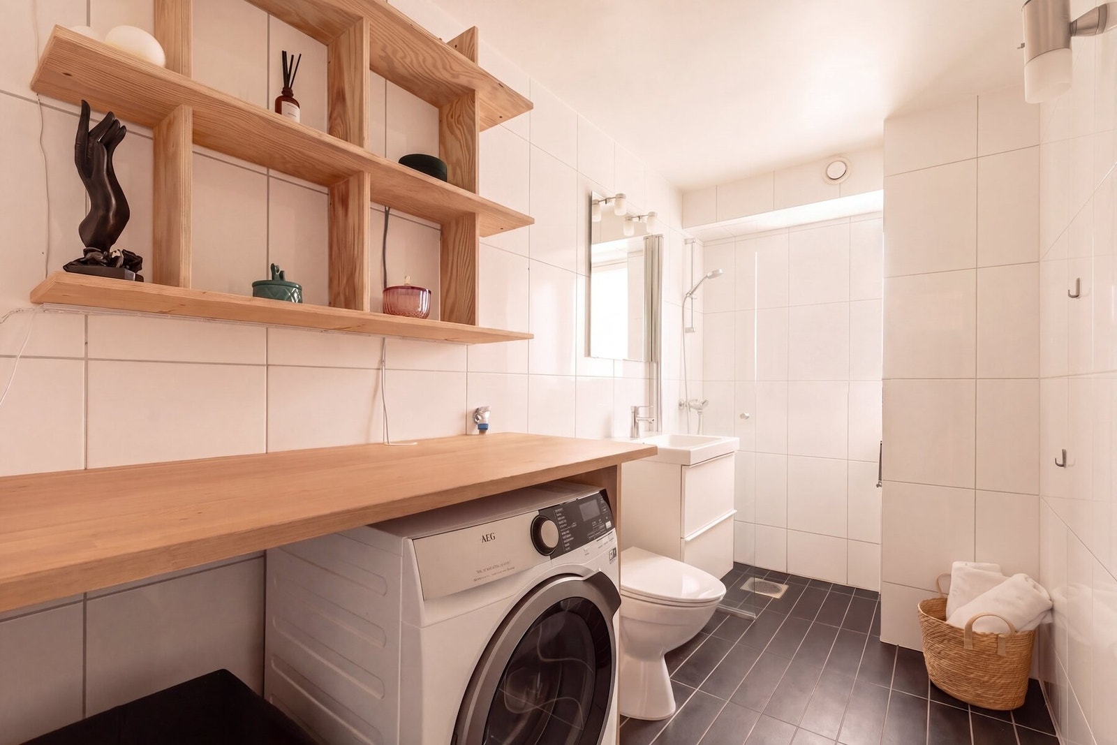 Badet har hvite veggfliser, sorte gulvfliser og opplegg for vaskemaskin - innbygd i en skreddersydd trebenk med åpne trehyller over. Galleribilde