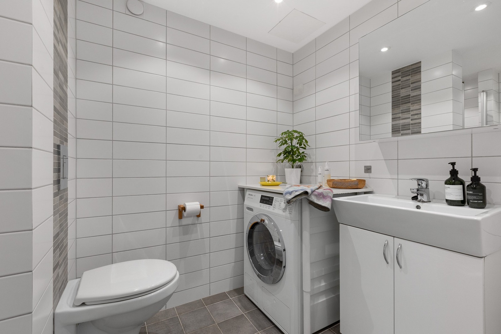 Badet inneholder dusjhjørne med glassvegg, vegghengt toalett, servant med underskap og plass til vaskemaskin. Galleribilde