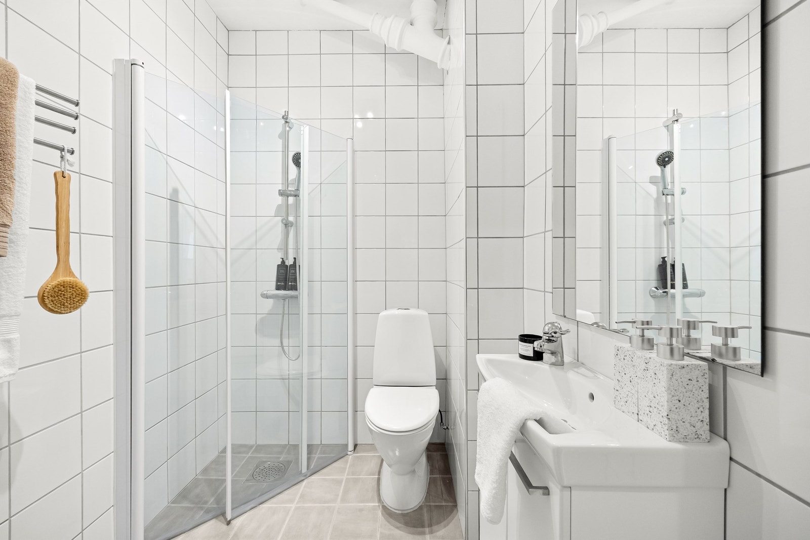 Badet er utstyrt med gulvstående toalett, vegghengt servantinnredning med ovenpåliggende vask og dusjnisje med dører. Galleribilde