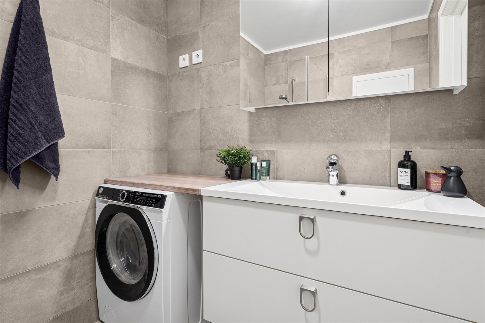 Badet er utstyrt med en stilren innredning med nedfelt servant, overskap med speil, veggmontert toalett, dusjhjørne og opplegg for vaskemaskin. Galleribilde