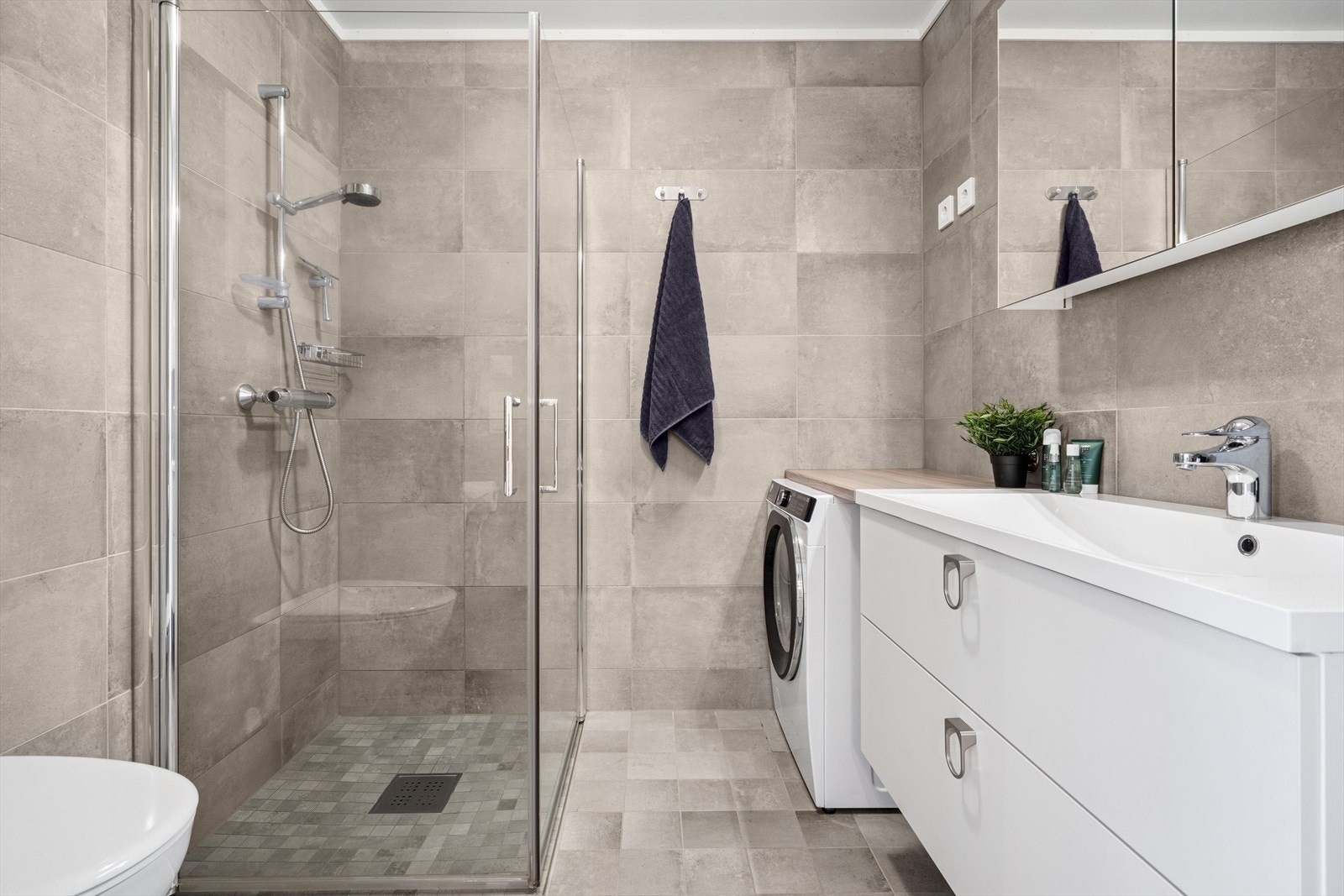 Det lyse og helflisede badet har elektriske varmekabler i gulvet for ekstra komfort. Badet er utstyrt med en stilren innredning med nedfelt servant, overskap med speil, veggmontert toalett og dusjhjørne. Galleribilde
