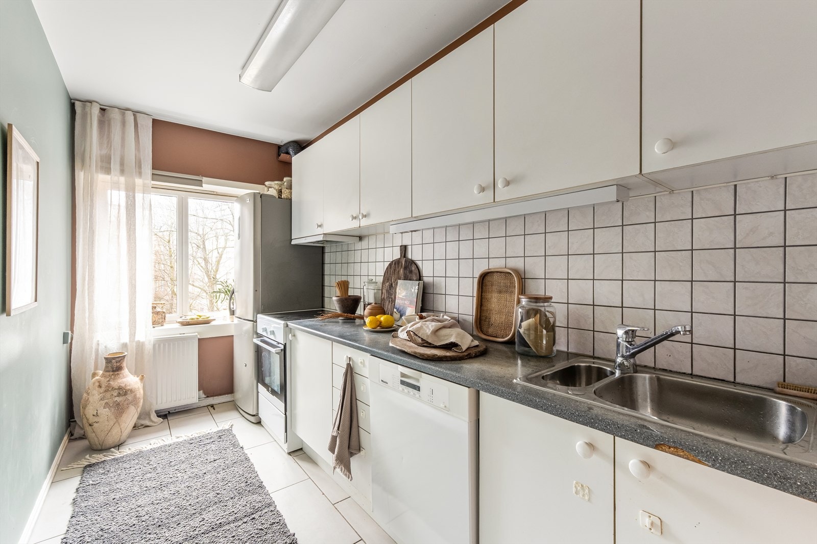 Eldre kjøkkeninnredning med slette fronter, fliser over laminat benkeplate med oppvaskkum og 1-greps blandebatteri. Det er ventilator med kullfilter over komfyr. Galleribilde
