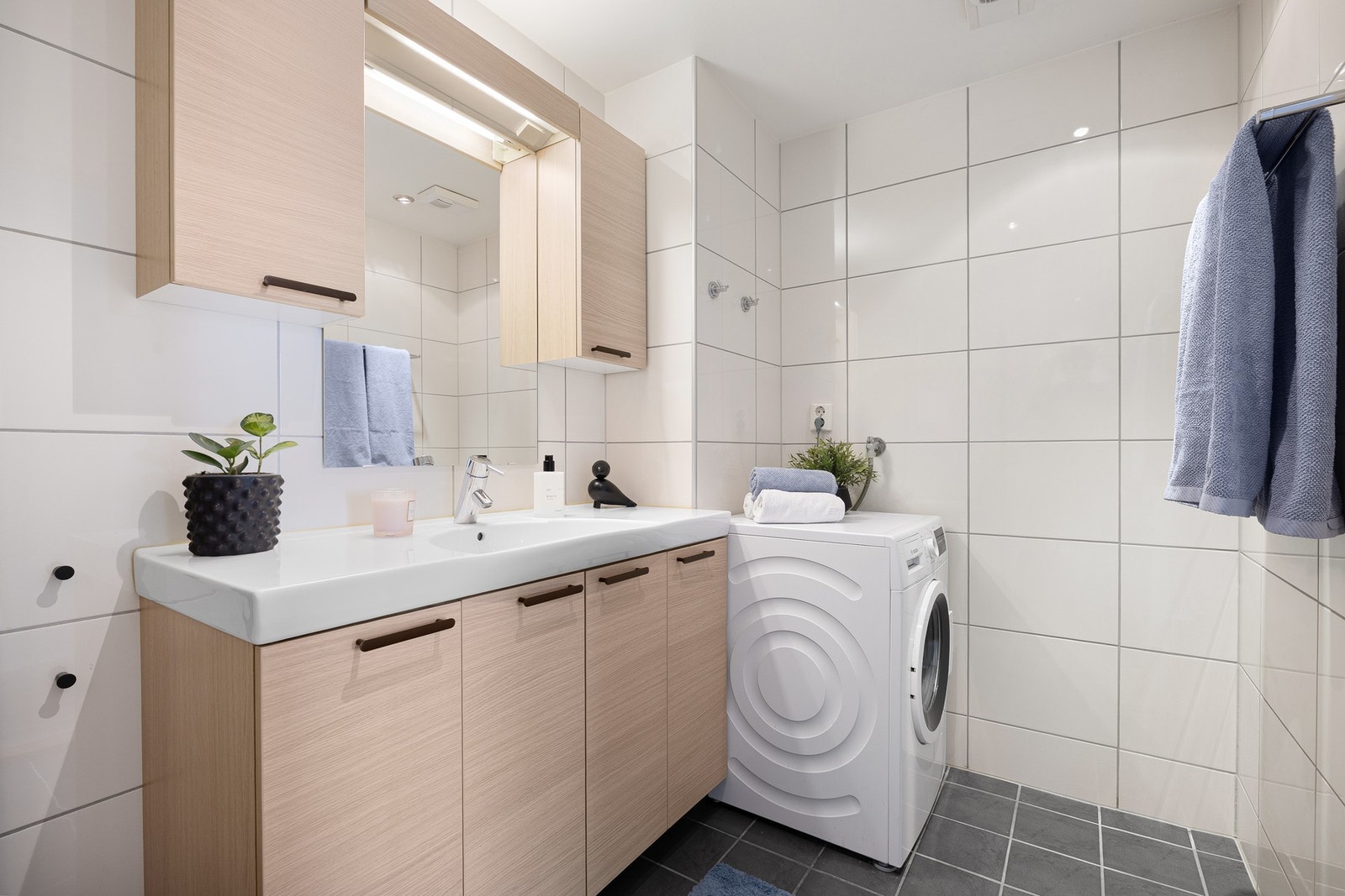 Badet inneholder. Innfellbare og plassbesparende dusjdører i glass, dusjbatteri og garnityr på vegg, veggmontert WC og kran og avløp for vaskemaskin. Nye dusjdører ble montert i 2024. Galleribilde