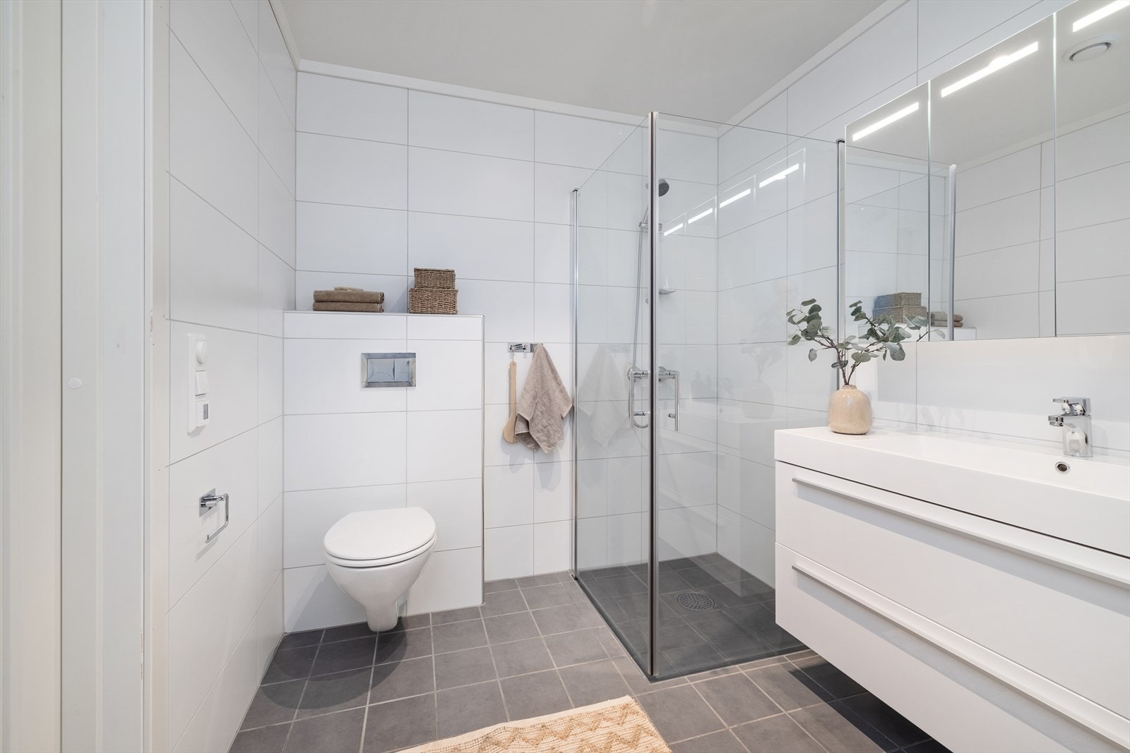 Badet er utstyrt med vegghengt toalett, servantinnredning med speilskap og et dusjhjørne med nedsenket sone. Galleribilde
