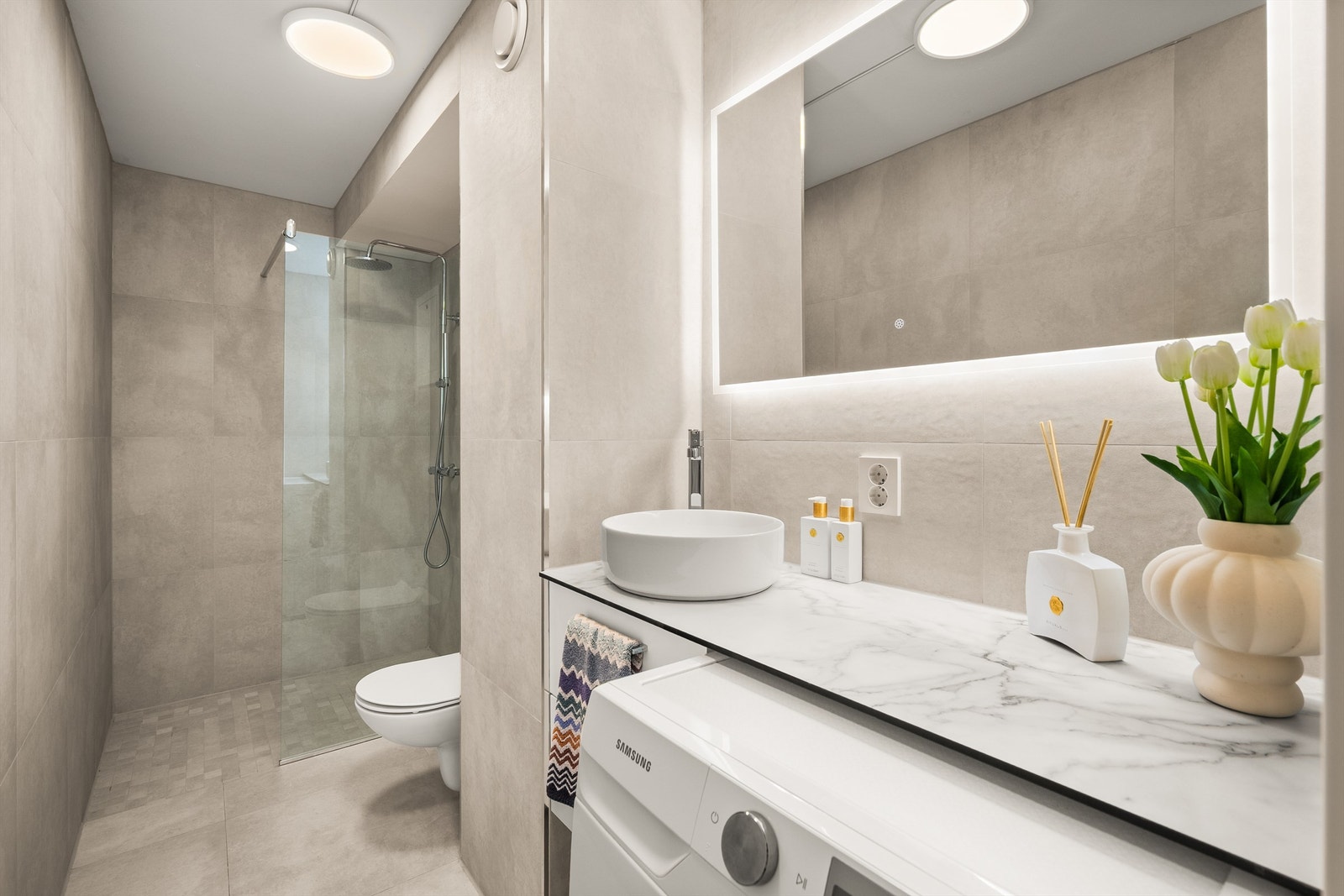 Lekkert, flislagt bad med gulvvarme fra 2025. Badet er innredet med vegghengt toalett, dusjhjørne med glassvegg og opplegg for vaskemaskin. Galleribilde