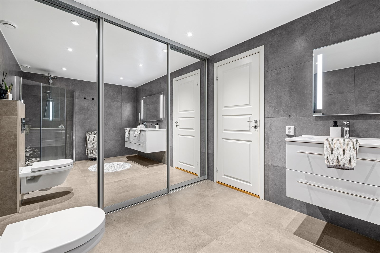 Badet er utstyrt med veggfestet toalett, dusjhjørne og en nedfelt servant med skuffeinnredning under. Bak en skyvedør med speilfronter er det opplegg for vaskemaskin. Galleribilde