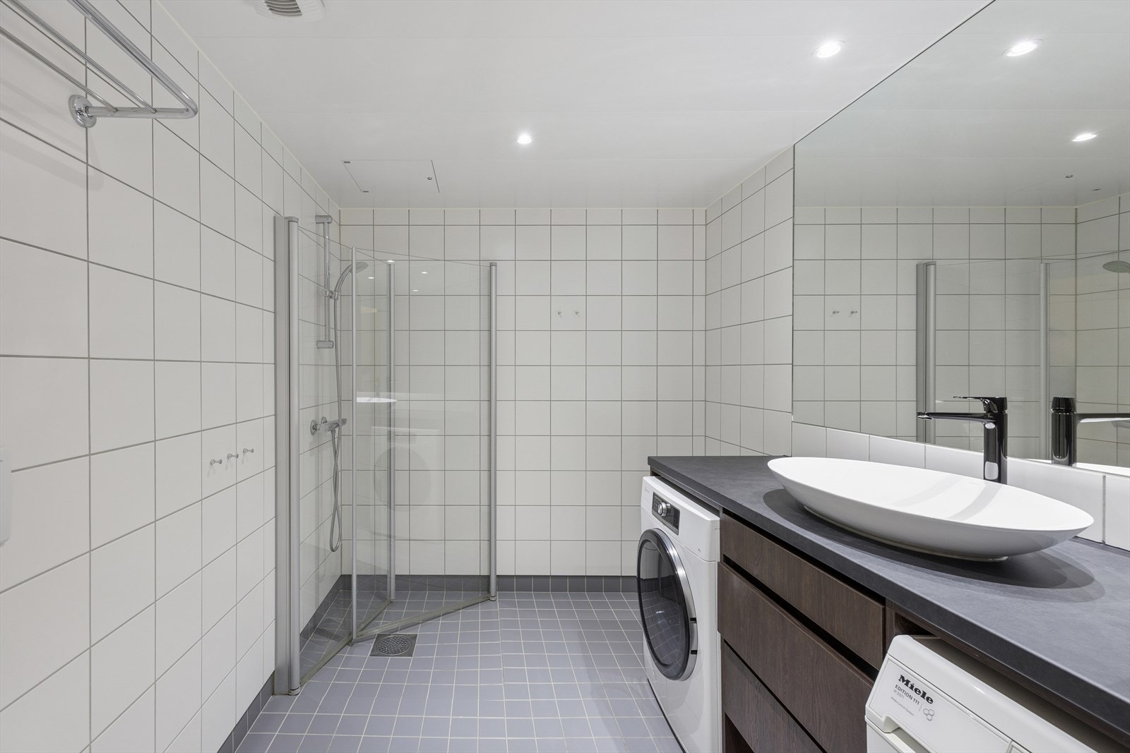 Badet har servantinnredning med skuffer, laminat benkeplate, nedfelt kum med ettgreps blandebatteri, speilskap, dusjhjørne med glassdører og vegghengt toalett. Galleribilde