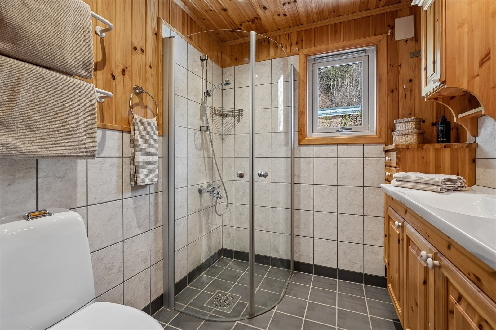 Badet er innredet med flislagt gulv med varmekabler, innredning i heltre furu med heldekkende servantplate, overskap, speil, gulvmontert toalett og dusj med dusjvegger. Galleribilde