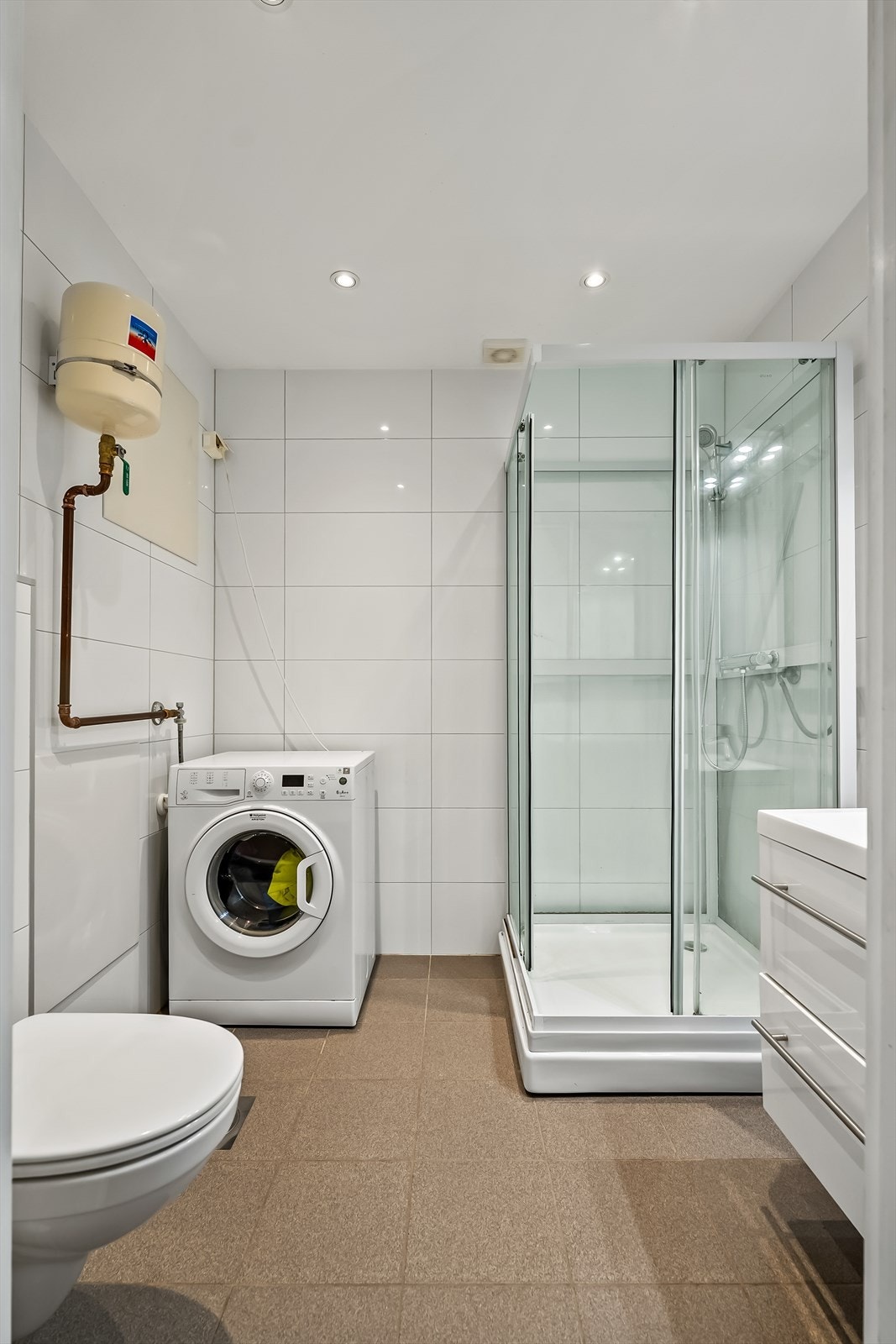 Badet er utstyrt med et veggmontert toalett, opplegg for vaskemaskin, dusjkabinett i herdet glass, speil med overliggende belysning, baderomsinnredning med høyglans fronter og heldekkende servant. Et moderne og funksjonelt rom. Galleribilde