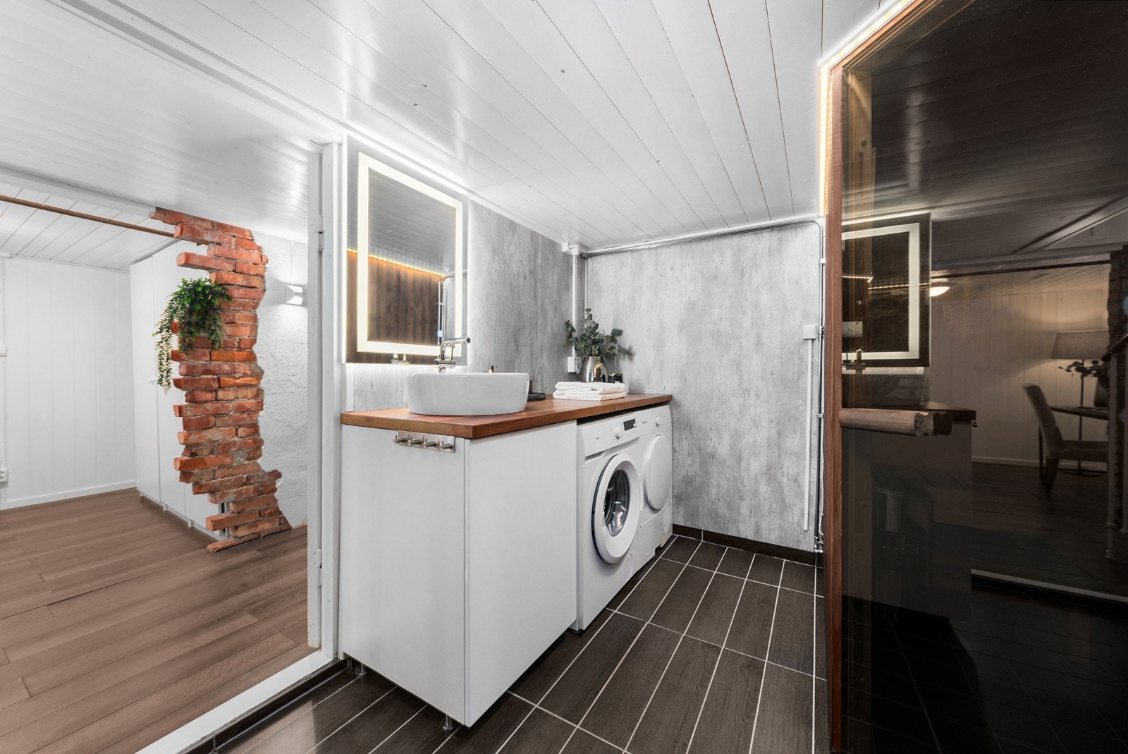 Et speil med integrert belysning er plassert over en hvit skapseksjon med stor benkeplate og toppmontert servantskål. Under benken har badet opplegg for vaskemaskin og plass til tørketrommel. Galleribilde