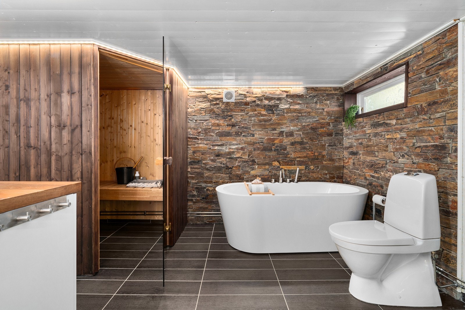 Badet nede har både en egen badstue, et dusjhjørne m/ innfellbare glassdører og et frittstående badekar i elegant design. På gulvet er det mørke fliser m/ varme. Veggene er kledd m/ grå våtromsplater, beiset trepanel og dekorfliser som ligner teglstein. Galleribilde