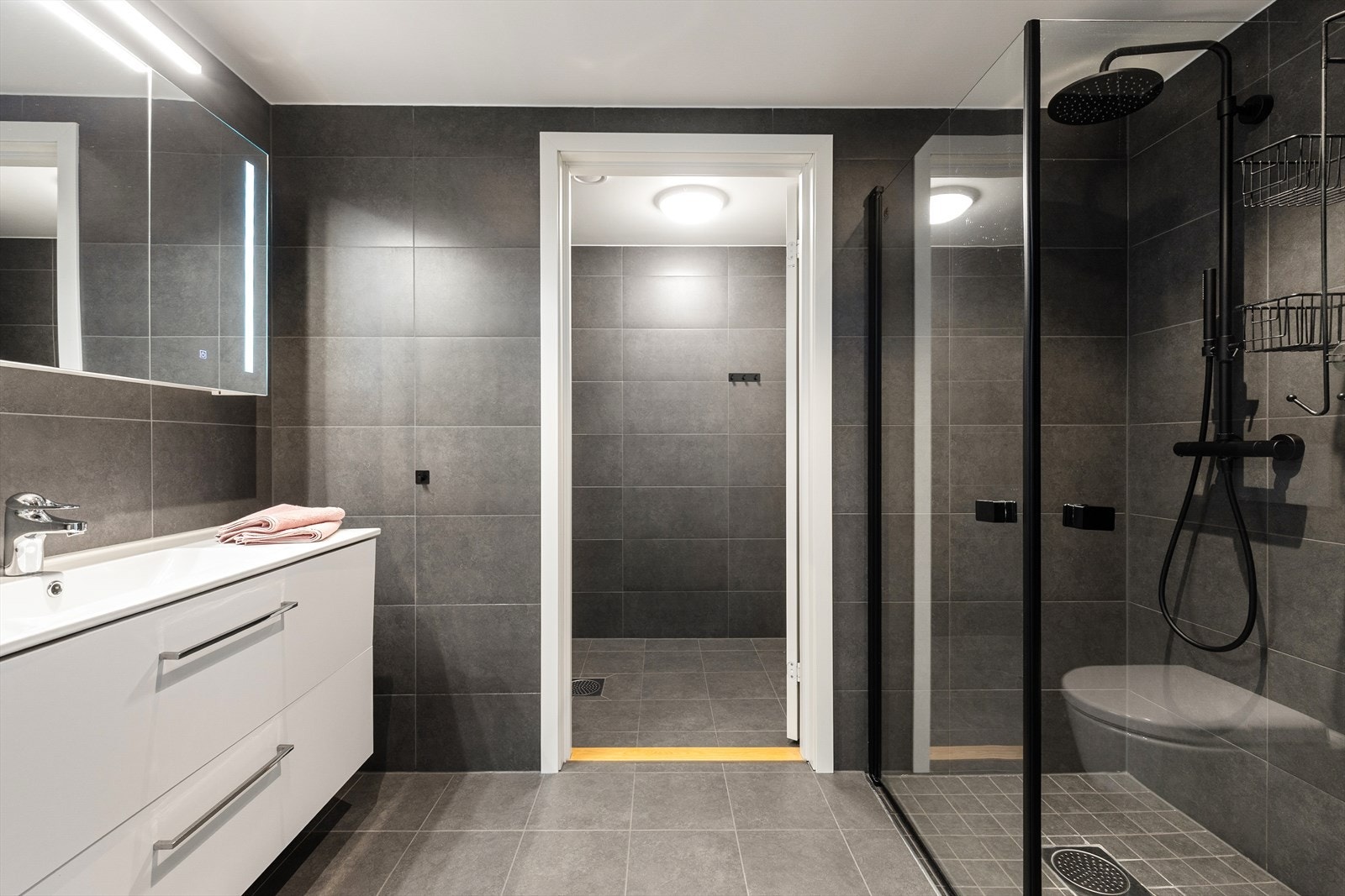 Flott bad med flis på gulv og vegger. Badet har vegghengt toalett, dushjørne med vegger og innredning med heldekkende hvit vask, underskuffer med slette fronter og overskap med speildører med lys. Det er varmekabler i gulv og balansert ventilasjon. Galleribilde