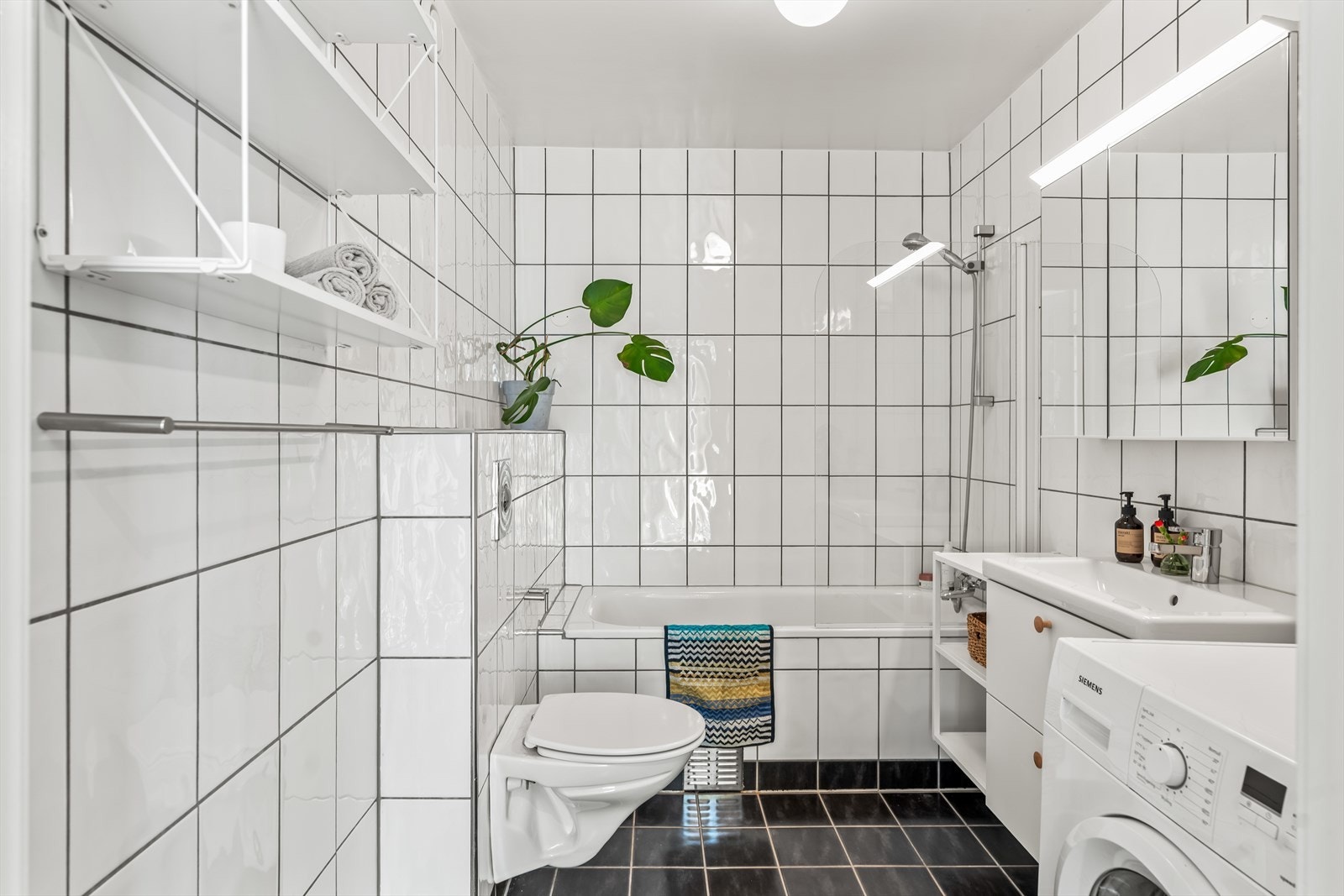 Badet inneholder speilinnredning med lys, vegghengt toalett og dusj/badekar bak glassvegg Galleribilde