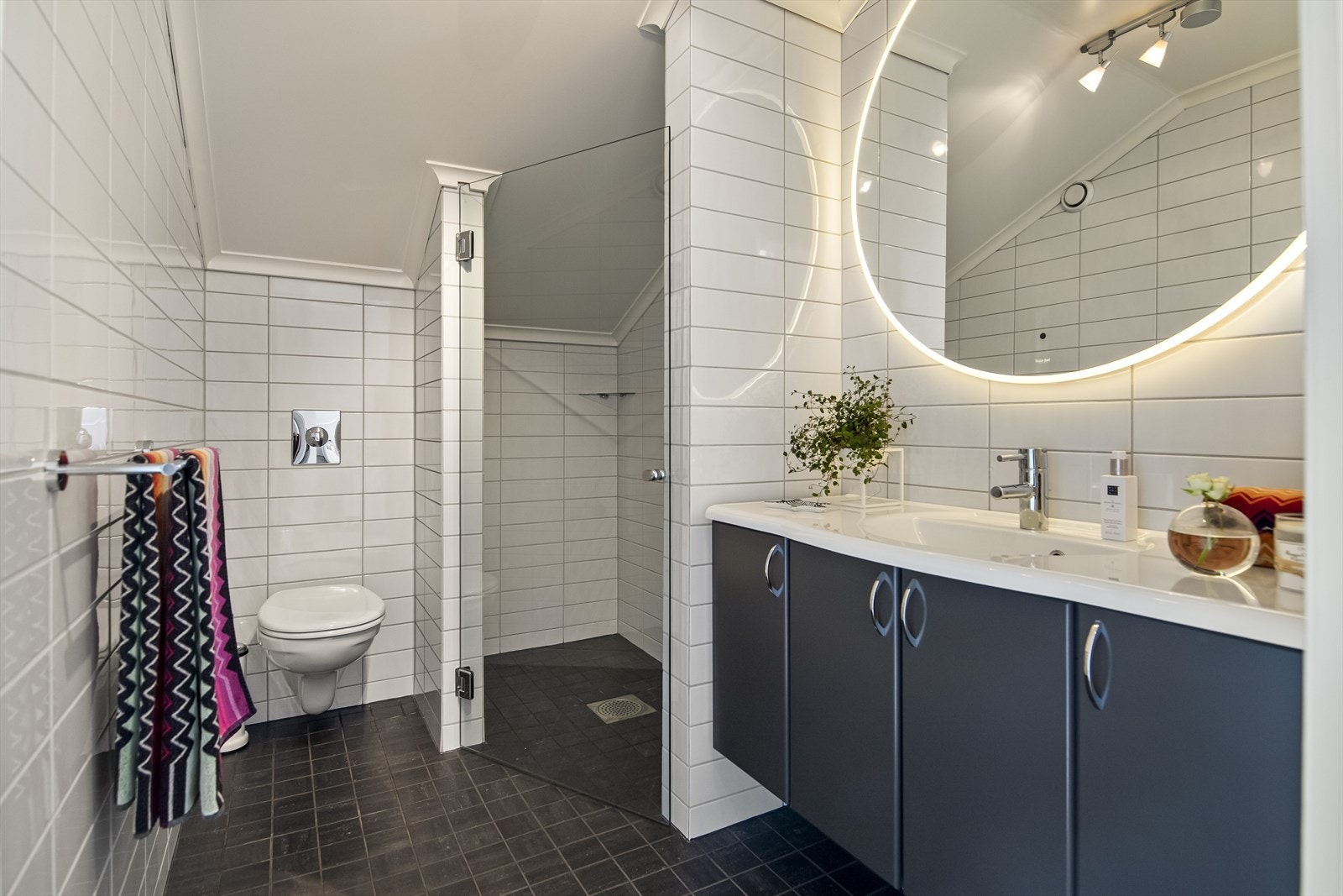 I loftsetasjen har badet mørkegrå gulvfliser og hvite veggfliser i ulike størrelser. På badet er det gulvvarme, vegghengt toalett og en dusjnisje med glassdør. Galleribilde