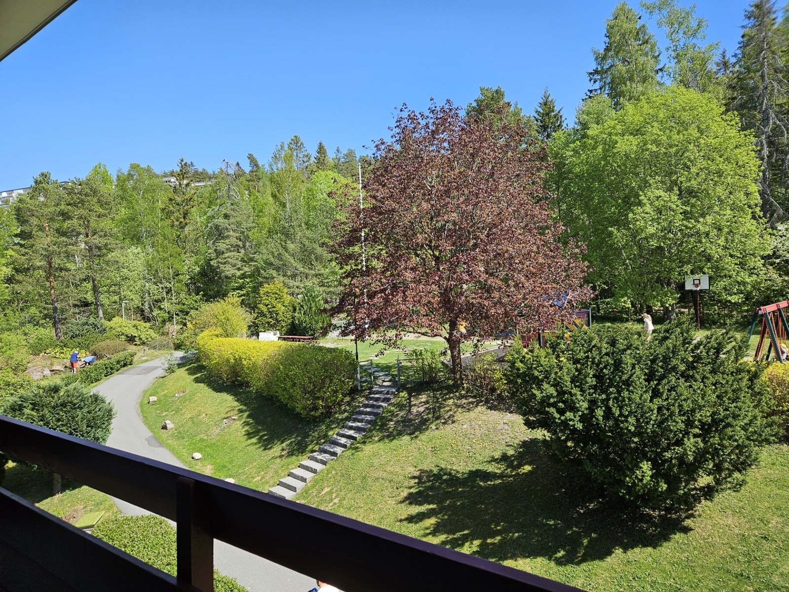 Fra balkongen er det et idyllisk utsyn over borettslagets grønne fellesområder - her ser du ned på en frodig og velholdt hage med gangsti, beplantning og lekeplass, med skogen som naturlig bakteppe. Galleribilde