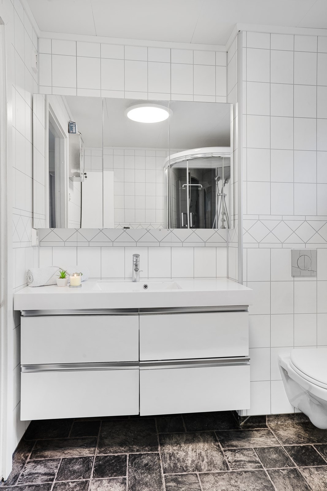 Innredningen på badet inkluderer underskap med vask, speilskap, veggmontert toalett og dusjkabinett. Galleribilde
