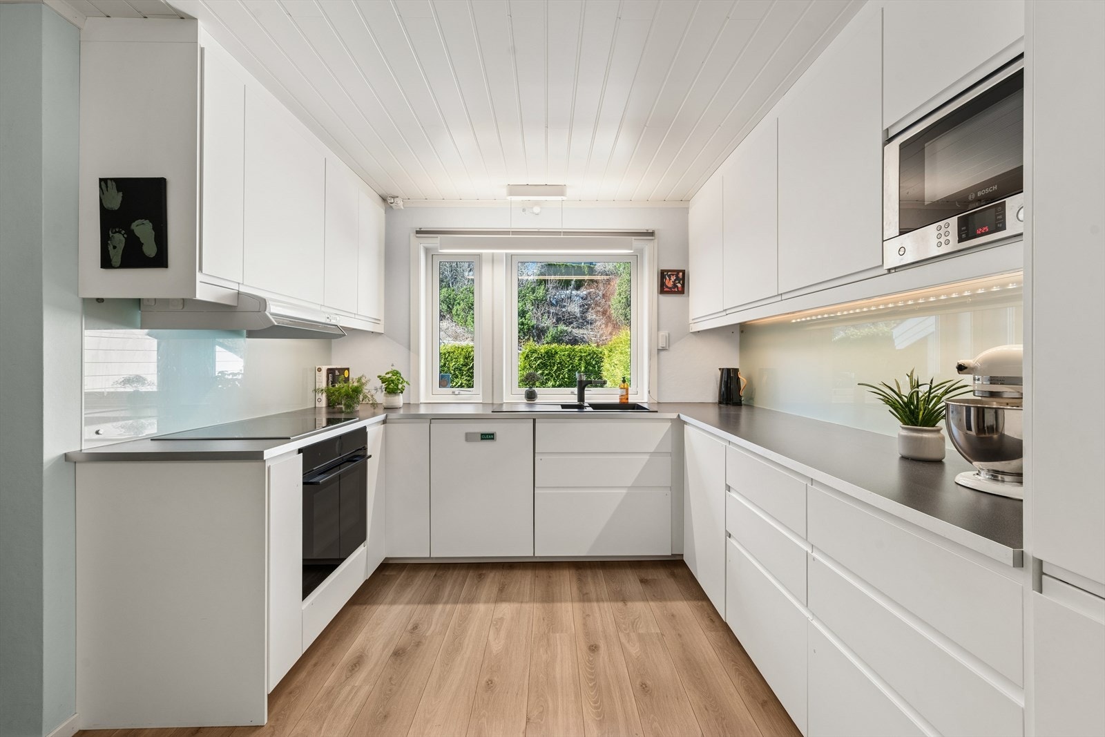 Nyere kjøkkeninnredning med bl.a. lyse glatte fronter, laminat benkeplate, glassplater over benken, ventilator med stål hette, sort kompositt benkebeslag og integrerte hvitevarer. Galleribilde