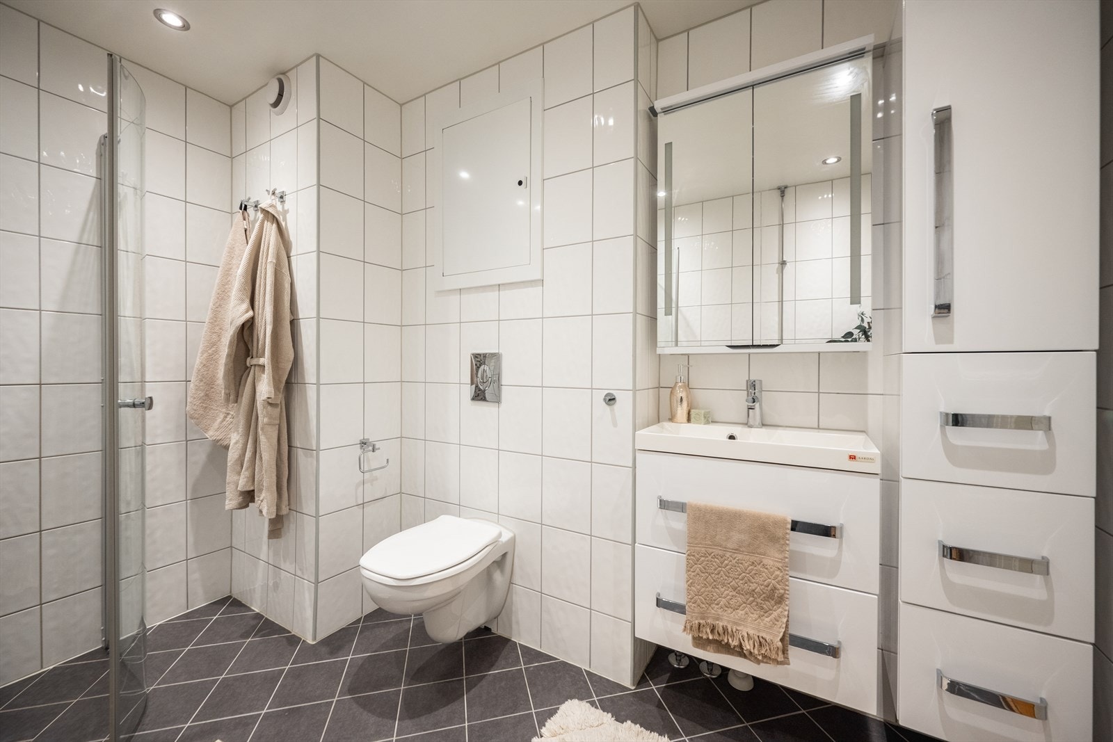 Badet er innredet med vegghengt klosett, servant med innredning, speilskap med lys og dusjhjørne med svingbare glassdører. Galleribilde