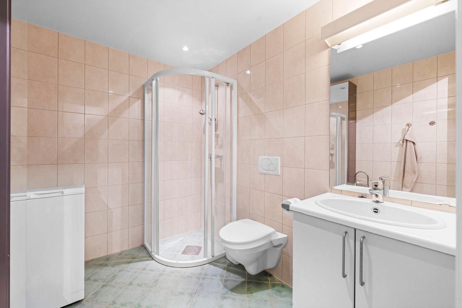 Flislagt bad med gulvvarme, servant på vegg med speil og belysning over. Badet har vegghengt toalett og dusjhjørne med glassdører. Galleribilde